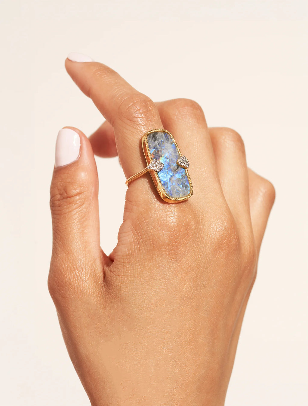 Hand wearing a gold ring with a large, faceted gemstone on a beige background