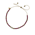 A beaded necklace with deep red faceted beads and metallic gold spacer beads, featuring an adjustable cord closure with a small gold leaf charm, laid flat on a light gray background.