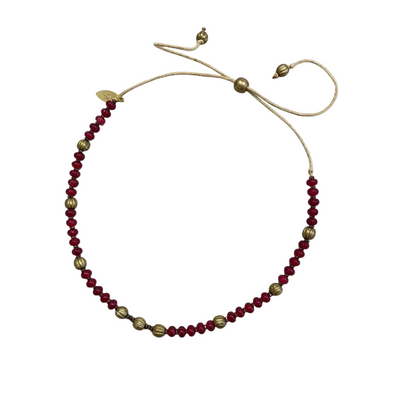 A beaded necklace with deep red faceted beads and metallic gold spacer beads, featuring an adjustable cord closure with a small gold leaf charm, laid flat on a light gray background.
