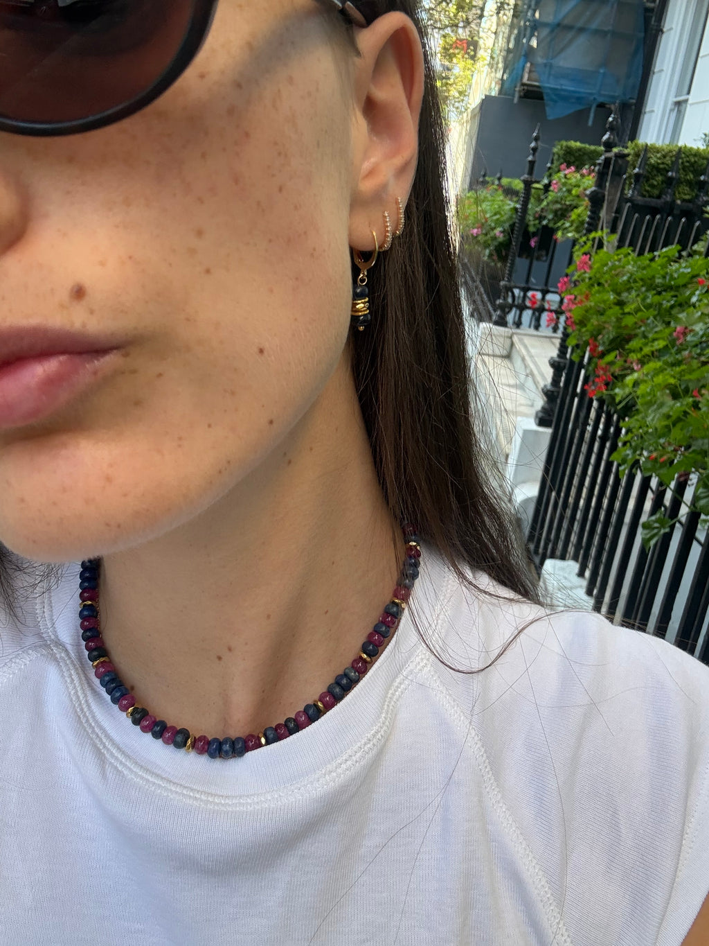 A close-up lifestyle shot of a person wearing a beaded necklace with dark red, black, and blue beads, along with gold hoop earrings, in an outdoor setting.