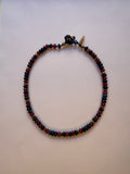 A beaded necklace with alternating dark blue and red beads, separated by small gold spacer beads, featuring a black and blue toggle clasp, displayed on a plain off-white background.